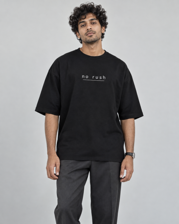 Model wearing a black oversized Toxikiing t-shirt with a small centred chest print reading 'no rush' in widely letter-spaced lowercase above a thin horizontal rule