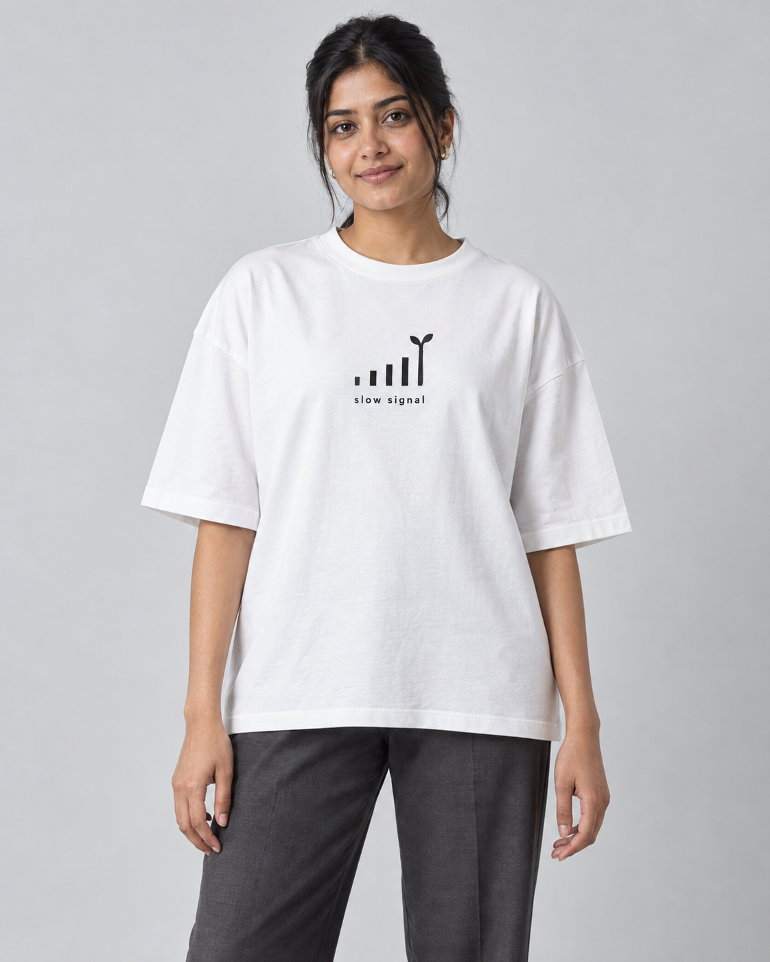 Model wearing a white oversized Toxikiing t-shirt with a small centred chest print of rising signal bars whose tallest bar becomes a sprouting seedling, captioned 'slow signal'
